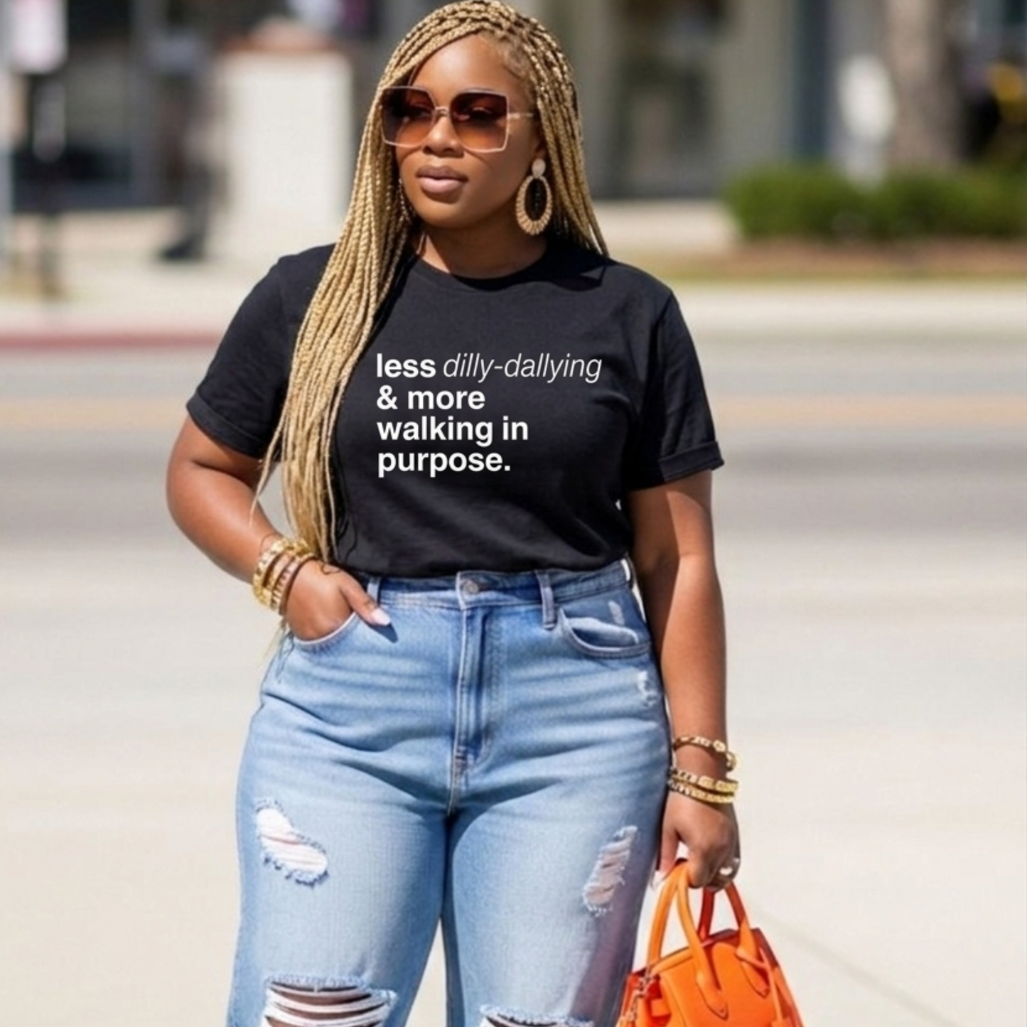 Woman wearing a black t-shirt with text, light blue jeans, and an orange handbag on a street.
